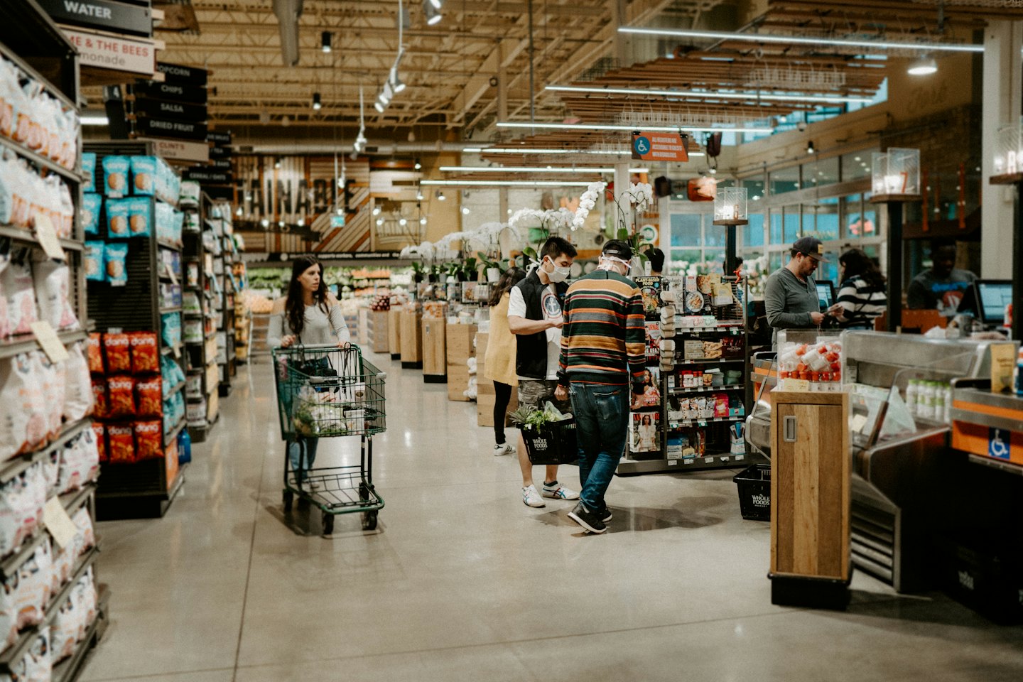 Einkäufer in einem modernen Supermarkt, wobei ein maskierter Mann mit jemandem spricht.